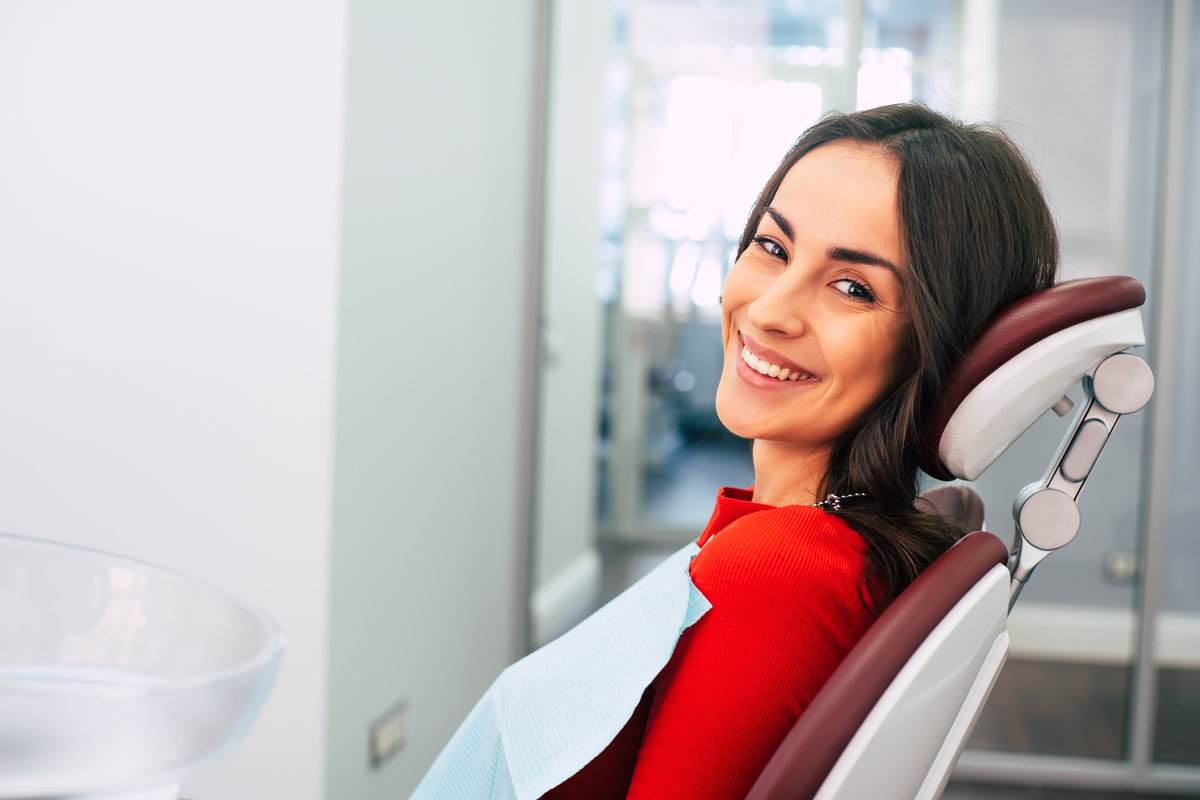 New life with new teeth. Gorgeous girl wearing red sweater in the stomatology room full of day-light and white colors is smiling with her new white eye-catching smile.
